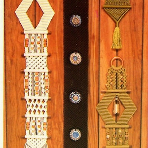 Può includere: Tre arazzi da parete in macramè con diversi design e colori. L'arazzo più a sinistra è bianco con perline marroni e una forma a diamante al centro. L'arazzo centrale è marrone con disegni circolari blu e bianchi. L'arazzo più a destra è verde con perline marroni e una campana in basso.