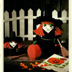 Może przedstawiać: Dwie szydełkowe lalki czarownic w czarnych kapeluszach i z czerwonymi włosami, szydełkowa pomarańczowa dynia, miotła i cukierki kukurydziane rozrzucone na białym tle z nadrukowaną serwetką z dynią.