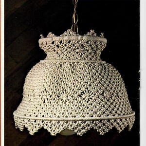 Op de afbeelding: Een witte macramé lampenkap met een delicaat, ingewikkeld patroon. De lampenkap hangt aan een ketting en heeft een witte gloeilamp erin.