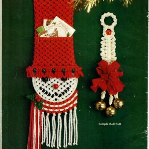 Puede incluir: Un porta tarjetas de crochet blanco y rojo con forma de muñeco de nieve con un borla rojo y blanco. El muñeco de nieve tiene una nariz roja y ojos negros. El porta tarjetas tiene un bolsillo para tarjetas. La imagen también muestra un tirador de campana de crochet blanco con un lazo rojo y tres campanas doradas.