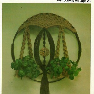 Peut inclure: Un porte-plantes mural en macramé avec trois anneaux et un pompon brun. Le porte-plantes est fait de fil brun et beige et a deux plantes vertes qui pendent de lui. Le texte "Three Ring Wall Planter" est en haut de l'image et "instructions on page 23" est en dessous.
