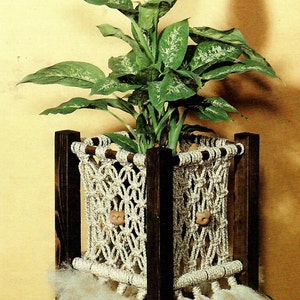 Op de afbeelding: Een macramé plantenhanger gemaakt van natuurlijke touw en houten pinnen. De hanger is 28 cm breed en 69 cm hoog. Hij kan een plantenpot met een diameter van 15 tot 20 cm bevatten. De hanger wordt getoond met een gepotte plant en een decoratieve pop.