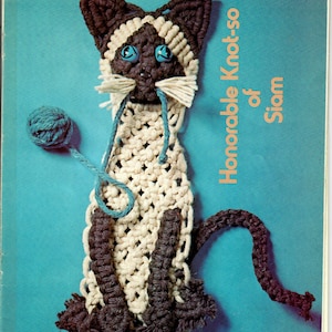 May include: A white and brown crocheted cat with blue eyes and a blue yarn ball. The cat is titled "Honorable Knot-so of Siam".