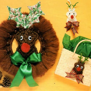 Puede incluir: Una corona de reno de lana marrón con un lazo verde, hojas de acebo verdes y una nariz roja de fieltro. También se muestran dos adornos de reno más pequeños, uno con un lazo a cuadros y el otro en una cesta tejida.