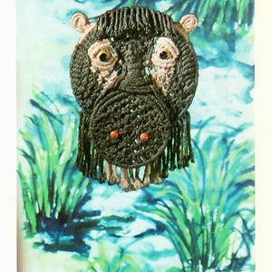 May include: A brown and black crocheted hippopotamus with pink ears and red beads for nostrils. The hippopotamus is hanging against a blue and green background with green leaves.