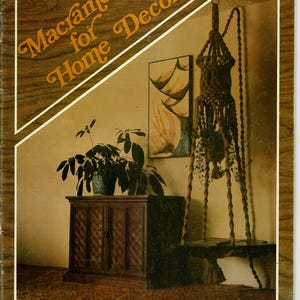 Könnte beinhalten: Ein Vintage-Buchcover mit dem Titel "Makramee für Heimdekoration" mit einem braunen Makramee-Pflanzenaufhänger, einem Holzschrank und einer Topfpflanze. Das Cover hat einen braunen Holzmaserungsrand und einen diagonalen weißen Streifen.