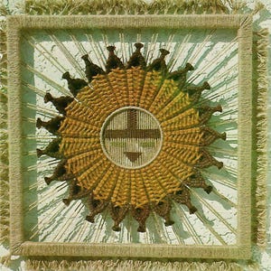 May include: A woven wall hanging with a brown and yellow circular design. The center of the design features a cross shape with a smaller triangle shape below it.