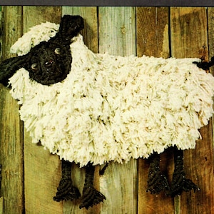 Könnte beinhalten: Ein weißes, flauschiges Schaf mit schwarzem Gesicht und Beinen. Das Schaf hängt an einer Holzwand.