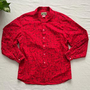 May include: A red button-down shirt with a black abstract pattern. The long-sleeved shirt has a collared neckline, a chest pocket, and white buttons. The shirt is made of a lightweight fabric.