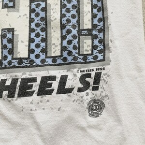May include: White t-shirt with a graphic design featuring the word "HEELS!" in large, distressed black letters. The design includes a blue and gray pattern at the top. The text "MR TEES 1995" is printed near the bottom.