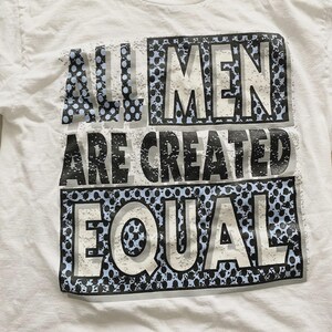 May include: White t-shirt with the text "ALL MEN ARE CREATED EQUAL" in a black, gray, and blue design. The text is arranged in three horizontal lines with a dotted pattern.