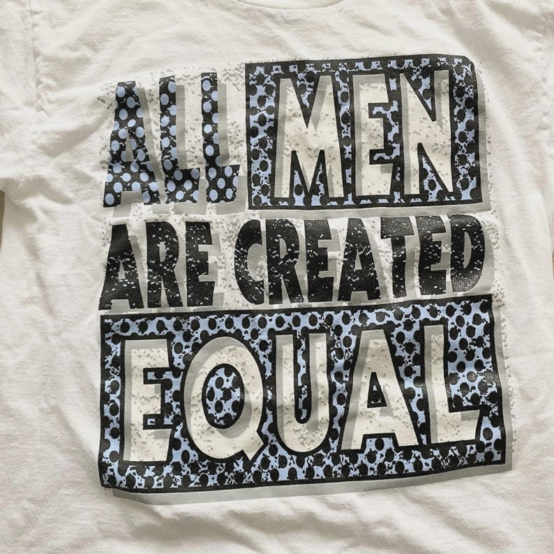May include: White t-shirt with the text "ALL MEN ARE CREATED EQUAL" in a black, gray, and blue design. The text is arranged in three horizontal lines with a dotted pattern.