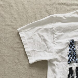 May include: White t-shirt with a graphic design on the front. The design includes the letters "A" and "M" in a black and blue dotted pattern. The shirt's sleeve is visible, showing the fabric's texture.
