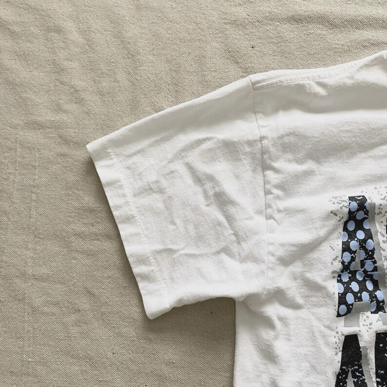 May include: White t-shirt with a graphic design on the front. The design includes the letters "A" and "M" in a black and blue dotted pattern. The shirt's sleeve is visible, showing the fabric's texture.