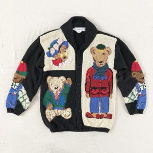 May include: A black cardigan sweater with a shawl collar and button closure. The sweater features colorful embroidered teddy bear designs on the front and sleeves. The bears are wearing hats, scarves, and outfits.