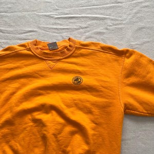 May include: Bright orange crewneck sweatshirt with a small circular logo on the chest. The logo features a globe design with text. The sweatshirt has a classic fit with ribbed cuffs and a crew neckline.