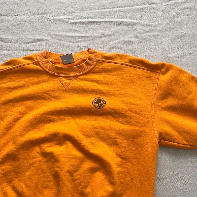 May include: Bright orange crewneck sweatshirt with a small circular logo on the chest. The logo features a globe design with text. The sweatshirt has a classic fit with ribbed cuffs and a crew neckline.