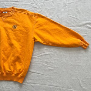 May include: Mustard yellow crew neck sweatshirt with a small embroidered emblem on the chest. The long sleeves have ribbed cuffs. The sweatshirt is made of a soft, comfortable material.