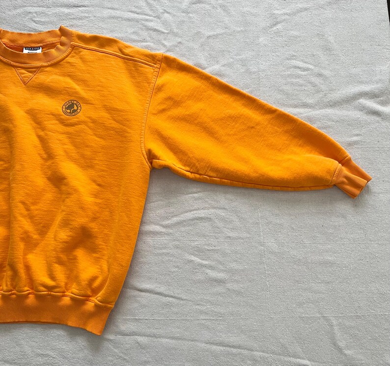 May include: Mustard yellow crew neck sweatshirt with a small embroidered emblem on the chest. The long sleeves have ribbed cuffs. The sweatshirt is made of a soft, comfortable material.