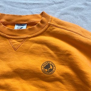 May include: Mustard yellow sweatshirt with a crew neck and the "Mossy Oak Camouflage" logo on the chest. The sweatshirt has a tag that reads "Jerzees". The garment is made of a soft material.