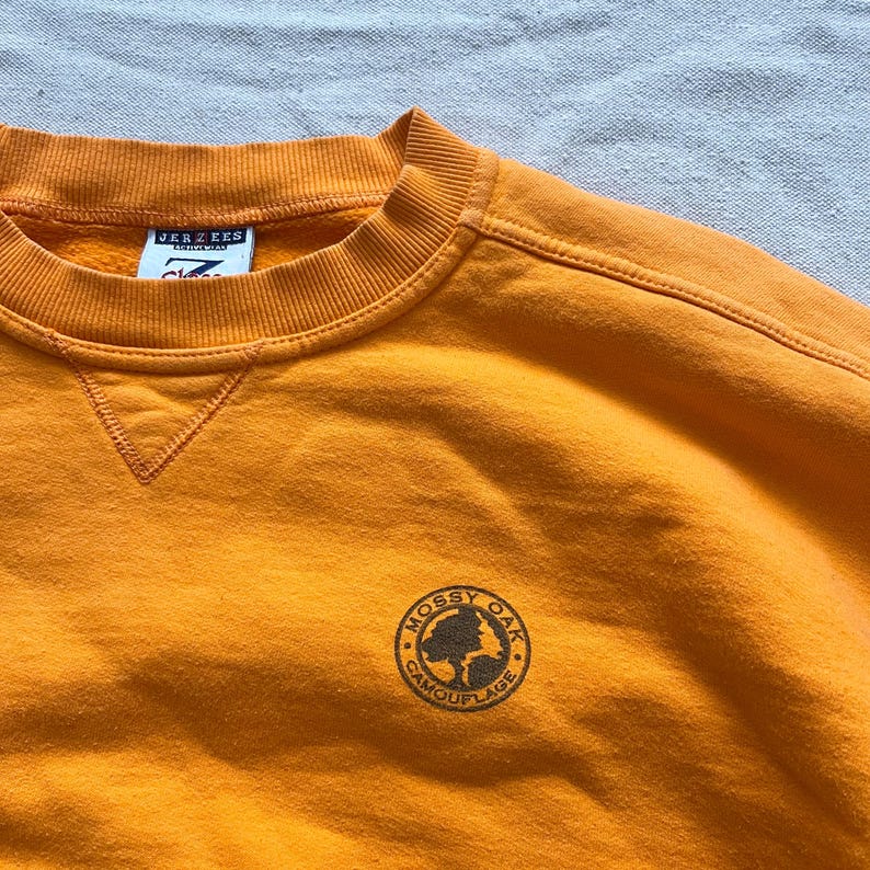 May include: Mustard yellow sweatshirt with a crew neck and the "Mossy Oak Camouflage" logo on the chest. The sweatshirt has a tag that reads "Jerzees". The garment is made of a soft material.