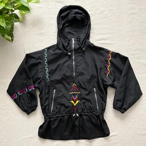 May include: Black hooded anorak jacket with colorful embroidered geometric patterns. Features a front zipper, two side pockets, and a drawstring waist. The jacket has a hood and long sleeves.