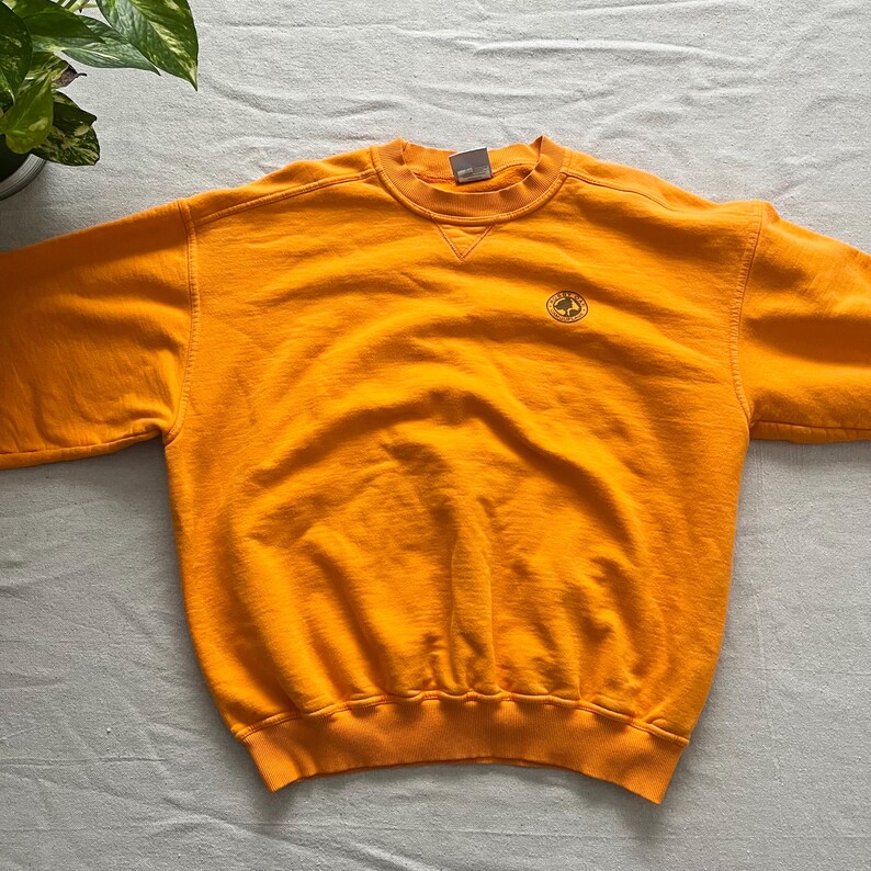 May include: Bright orange crewneck sweatshirt with a small circular emblem on the chest. The sweatshirt has a classic design with a ribbed collar, cuffs, and hem. The fabric appears soft and comfortable, suitable for casual wear.