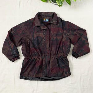 May include: A vintage jacket with a colorful geometric pattern in shades of burgundy, brown, and navy blue. The jacket features a stand-up collar, a drawstring waist, and a black bottom hem. The jacket has a button and zipper closure.