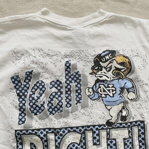 May include: White t-shirt featuring a cartoon ram mascot in a light blue sweater and a black cap with "UNC" on it. The shirt has the words "Yeah Right!" in a blue and black dotted design.