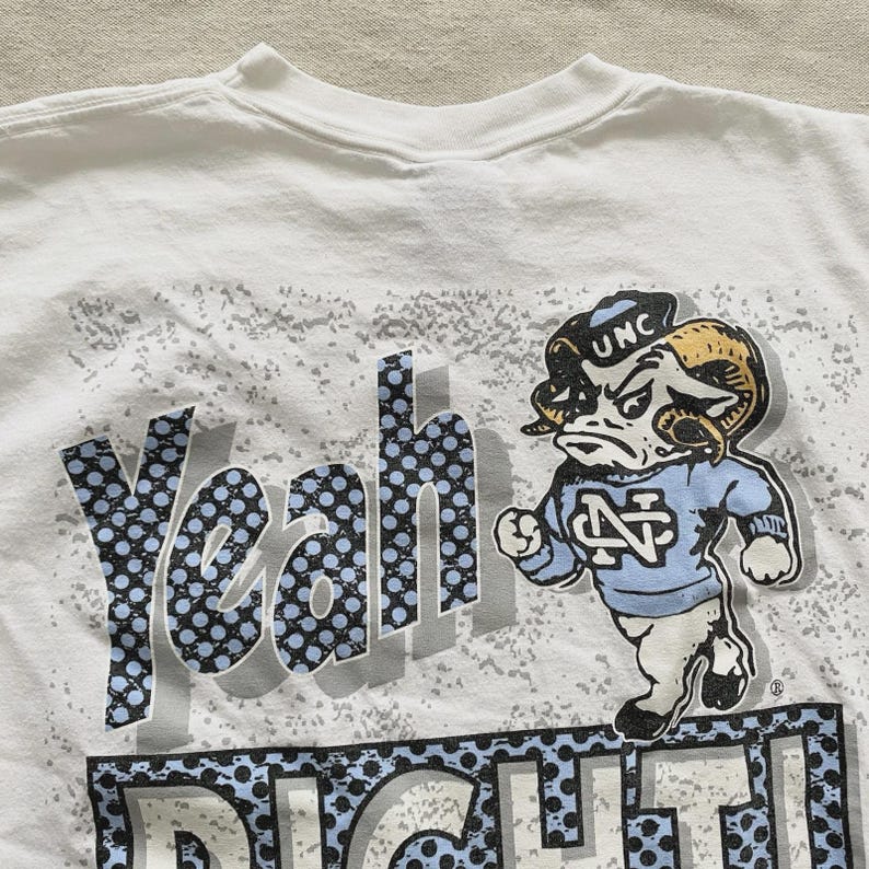 May include: White t-shirt featuring a cartoon ram mascot in a light blue sweater and a black cap with "UNC" on it. The shirt has the words "Yeah Right!" in a blue and black dotted design.