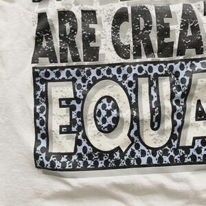 May include: White t-shirt with the text "ARE CREATED EQUAL" in a distressed, textured font. The word "EQUAL" is in a white font with a blue and black dotted border. The shirt is made of a soft, white fabric.