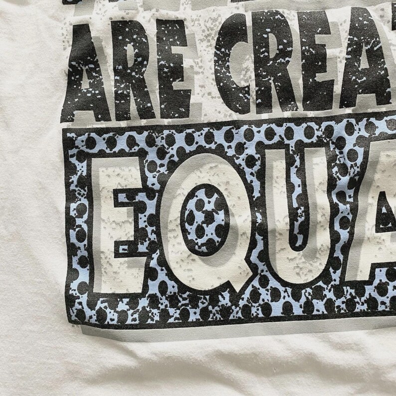 May include: White t-shirt with the text "ARE CREATED EQUAL" in a distressed, textured font. The word "EQUAL" is in a white font with a blue and black dotted border. The shirt is made of a soft, white fabric.