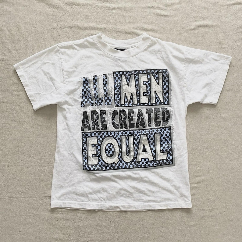 May include: White t-shirt with the text "ALL MEN ARE CREATED EQUAL" in a black and blue design. The shirt is short-sleeved and has a crew neck. The text is in a bold, blocky font.
