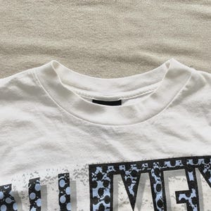 May include: White crew neck t-shirt with a graphic design. The graphic features the word "MEN" in a stylized font with blue and black accents. The shirt is made of a soft, comfortable material.