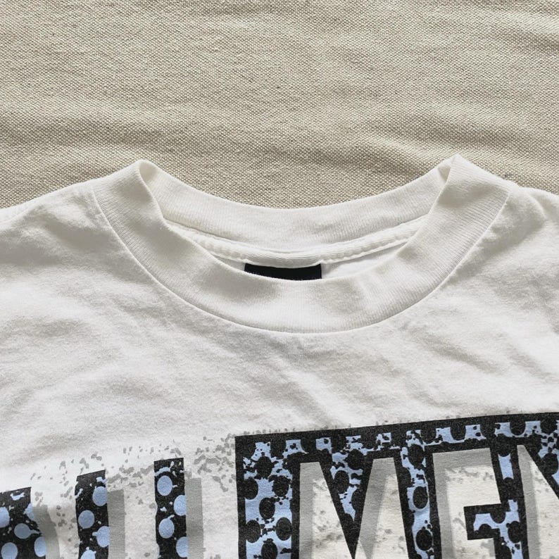 May include: White crew neck t-shirt with a graphic design. The graphic features the word "MEN" in a stylized font with blue and black accents. The shirt is made of a soft, comfortable material.