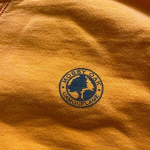 May include: Close-up of a mustard-colored fabric with a circular navy blue logo. The logo reads "Mossy Oak Camouflage" around the edge, with a tree graphic in the center. The fabric has a slightly textured appearance.