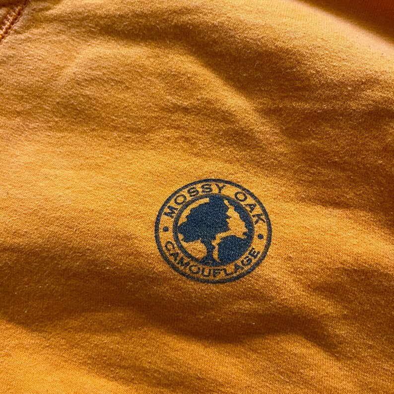 May include: Close-up of a mustard-colored fabric with a circular navy blue logo. The logo reads "Mossy Oak Camouflage" around the edge, with a tree graphic in the center. The fabric has a slightly textured appearance.