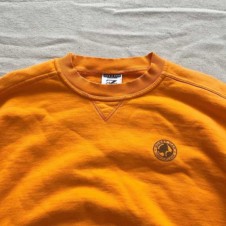 May include: Mustard-colored sweatshirt with a crew neck and a small embroidered logo on the chest. The logo is a circular design with the words "Mossy Oak Camouflage" around a tree graphic. The sweatshirt has a tag that reads "JERZEES".