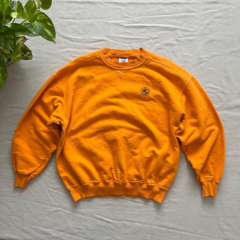 May include: Bright orange sweatshirt with a crew neck and long sleeves. A small circular emblem is on the upper left chest. The sweatshirt is made of a soft material.