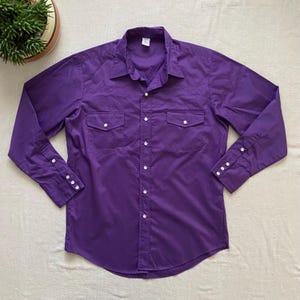 May include: A long-sleeved, solid purple button-down shirt with two chest pockets and white buttons. The shirt has a classic Western style with pointed front and back yokes. The cuffs also have white buttons.
