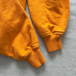 May include: Close-up of two orange sweatshirt sleeves. The cuffs are ribbed and a slightly darker shade of orange. The fabric has a textured appearance. The sleeves are lying on a light gray surface.