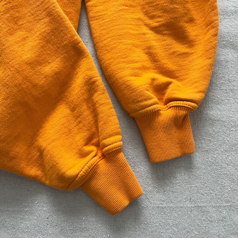 May include: Close-up of two orange sweatshirt sleeves. The cuffs are ribbed and a slightly darker shade of orange. The fabric has a textured appearance. The sleeves are lying on a light gray surface.