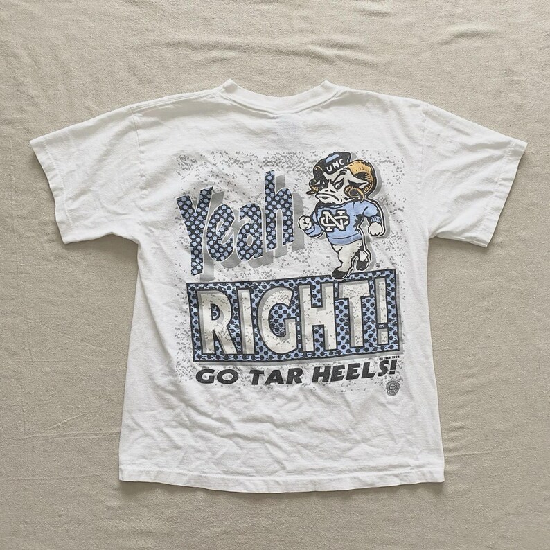 May include: White t-shirt with a graphic design featuring the words "Yeah RIGHT! GO TAR HEELS!" and a cartoon ram wearing a UNC hat and jersey. The design incorporates blue and gray accents.