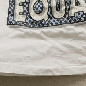 May include: White t-shirt with the word "EQUA" printed across the top in a distressed style. The letters are white with a blue and black speckled border. The shirt is made of a soft, white fabric.