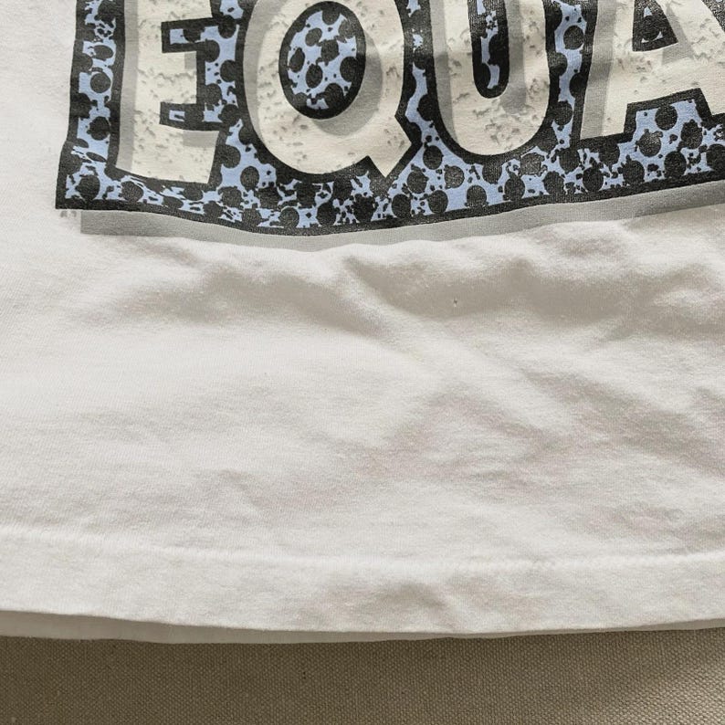 May include: White t-shirt with the word "EQUA" printed across the top in a distressed style. The letters are white with a blue and black speckled border. The shirt is made of a soft, white fabric.
