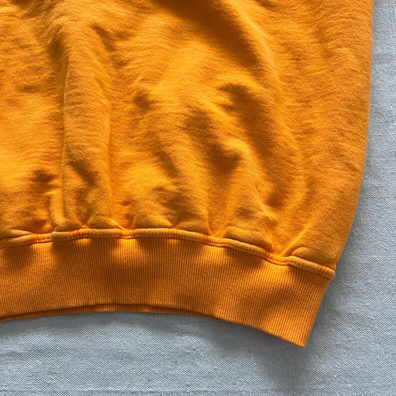 May include: Close-up of a bright orange sweatshirt. The ribbed hem and cuffs are a slightly darker shade of orange. The fabric has a soft, textured appearance, with subtle variations in color and shadow.