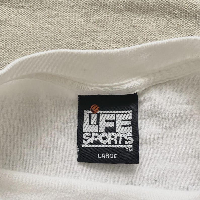 May include: Close-up of a white t-shirt with a black tag. The tag features the text "LIFE SPORTS" in white block letters, a basketball graphic, and the size "LARGE". The shirt is made of a soft, likely cotton material.