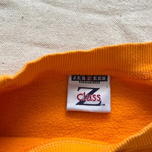 May include: Close-up of a bright orange sweatshirt with a ribbed neckline. A rectangular tag is sewn on the garment, displaying the text "JERZEES ACTIVEWEAR" above the word "Class" in a stylized font.