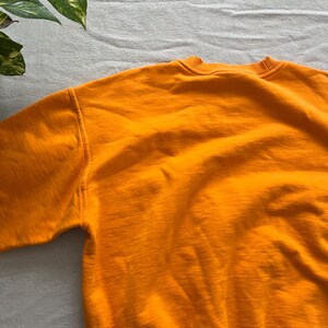 May include: A close-up of a bright orange sweatshirt. The crew neck and shoulder seams are visible. The fabric has a soft, textured appearance. The sweatshirt is made of a thick material, suitable for cooler weather.