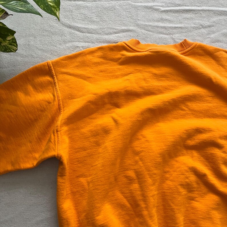 May include: A close-up of a bright orange sweatshirt. The crew neck and shoulder seams are visible. The fabric has a soft, textured appearance. The sweatshirt is made of a thick material, suitable for cooler weather.
